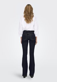 Jeans flare in denim scuro a vita alta con tasche posteriori e cuciture a contrasto, abbinati a una camicia bianca a maniche lunghe dalla vestibilità ampia.