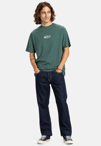 Zelené bavlnené tričko s malým logom na hrudníku, v kombinácii s tmavomodrými džínsami a čiernymi teniskami, stojí v uvoľnenej pôze.