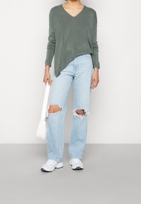 Grüner V-Ausschnitt-Pullover, hellblaue, ausgewaschene, weit geschnittene Jeans mit Rissen an den Knien, weiße Umhängetasche und weiße Sneakers mit schwarzen Akzenten.