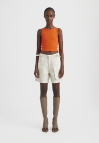 Top corto a coste arancione, pantaloni corti beige con tasche laterali e stivali al ginocchio marroni con tacco. Silhouette semplice e contemporanea con dettagli minimi.
