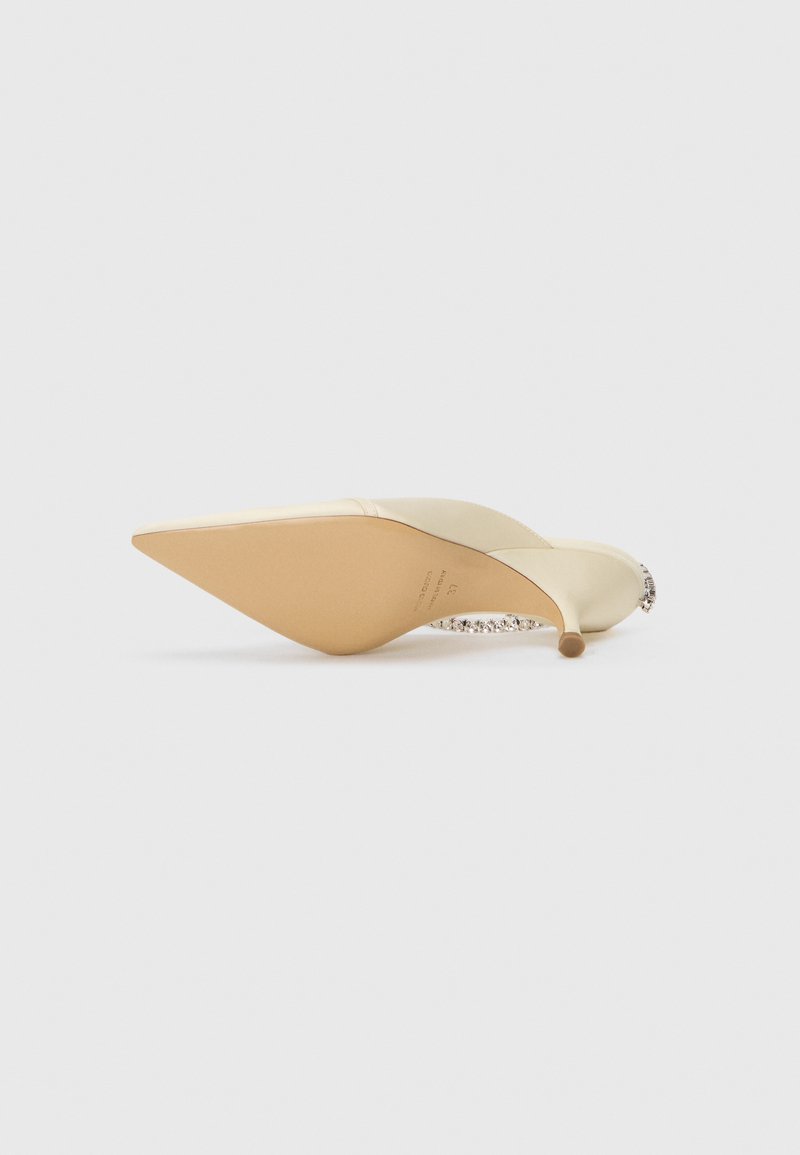 Spidse beige satin højhælede sko med en juvelbesat ankelrem, liggende på siden på en hvid baggrund.