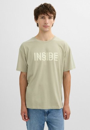 Joven con cabello corto y castaño, vistiendo una camiseta beige con el texto "INSIDE" y jeans azules, de pie frente a un fondo liso de color gris claro.