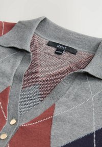 Cardigan gris à col avec motif losanges en rouge, gris et bleu marine, boutons dorés et étiquette de marque "NEXT" à l'intérieur du col.