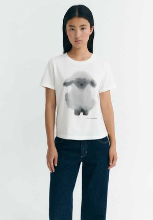 Junge Frau mit langen dunklen Haaren, die ein weißes T-Shirt mit einem grauen Aquarell-Schafdruck und dunkelblauen Jeans trägt, steht vor einem weißen Hintergrund.