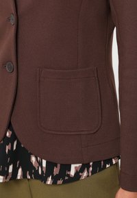 Brauner, strukturiert Blazer mit Knopfverschluss und einer genähten Tasche. Darüber getragen wird ein gemustertes Hemd und ein grüner Stoff, der feine Muster aufweist.