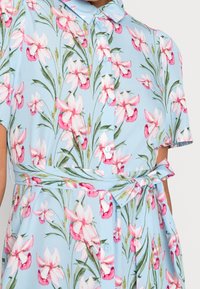 Robe florale en tissu léger, avec un fond bleu clair, des motifs de fleurs roses et blanches, manches courtes et taille ceinturée.