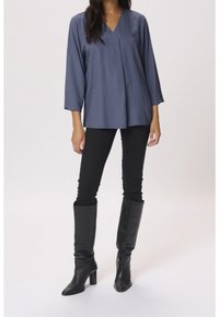 Femme portant un chemisier bleu texturé à manches longues, un pantalon noir slim et des bottes noires à talons montant jusqu'aux genoux, sur un fond uni.