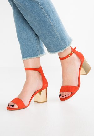 Rode suède hoge hak sandalen hebben een open teenontwerp, een enkelband met een gesp en een gouden blokhak. Draag ze met denim met een rafelrand.