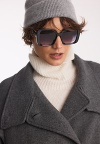 Czarne okulary przeciwsłoneczne z gradientowymi soczewkami, szary wełniany płaszcz oraz kremowy golf w prążki. Szara dziana czapka dopełnia całość.