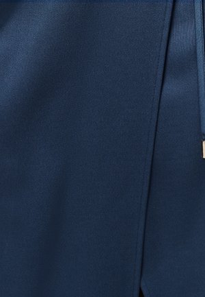 Primo piano di tessuto blu navy con una cucitura verticale e una vista parziale di una cinghia in tessuto abbinato con una estremità metallica.