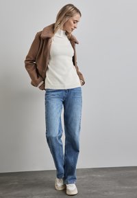 Bruine wollen jas met een schapenvachtkraag, witte geribde coltruien, blauwe recht gesneden jeans en witte sneakers.