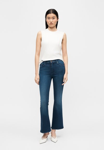 Γυναίκα που στέκεται μπροστά σε λευκό φόντο, φοράει λευκό αμάνικο τοπ, σκούρο μπλε φαρδιά τζιν και άσπρες μυτερές γόβες.