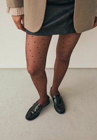 Zwarte polka dot maillot met een doorschijnende afwerking, gedragen met een grijze blazer en zwarte loafers op een lichtgekleurde vloer.