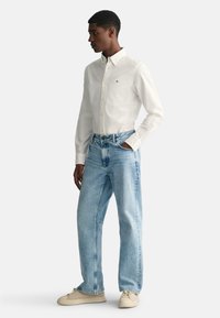 Chemise blanche à boutons, jeans baggy bleu clair et baskets beiges. La chemise arbore un petit logo ; les jeans possèdent un design classique à cinq poches.