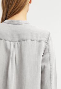 Camicia leggera grigia con dettaglio a pieghe sul retro e colletto classico. Realizzata in tessuto morbido e traspirante con una texture sottile.