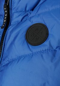 Plava puffer jakna s sjajnom završnom obradom, horizontalnim šavovima i okruglom crnom gumenom etiketom s utisnutim natpisom 'NMT 97'. Zatvaranje na zip.