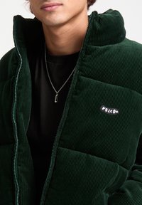Green corduroy puffer jacket with zip closure, featuring a high collar and a black logo patch. Worn over a black shirt, showing a silver necklace.