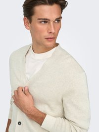 Cardigan beige clair à col en V, tricot texturé et manches longues, avec un seul bouton foncé sur le devant, porté par-dessus une chemise blanche.