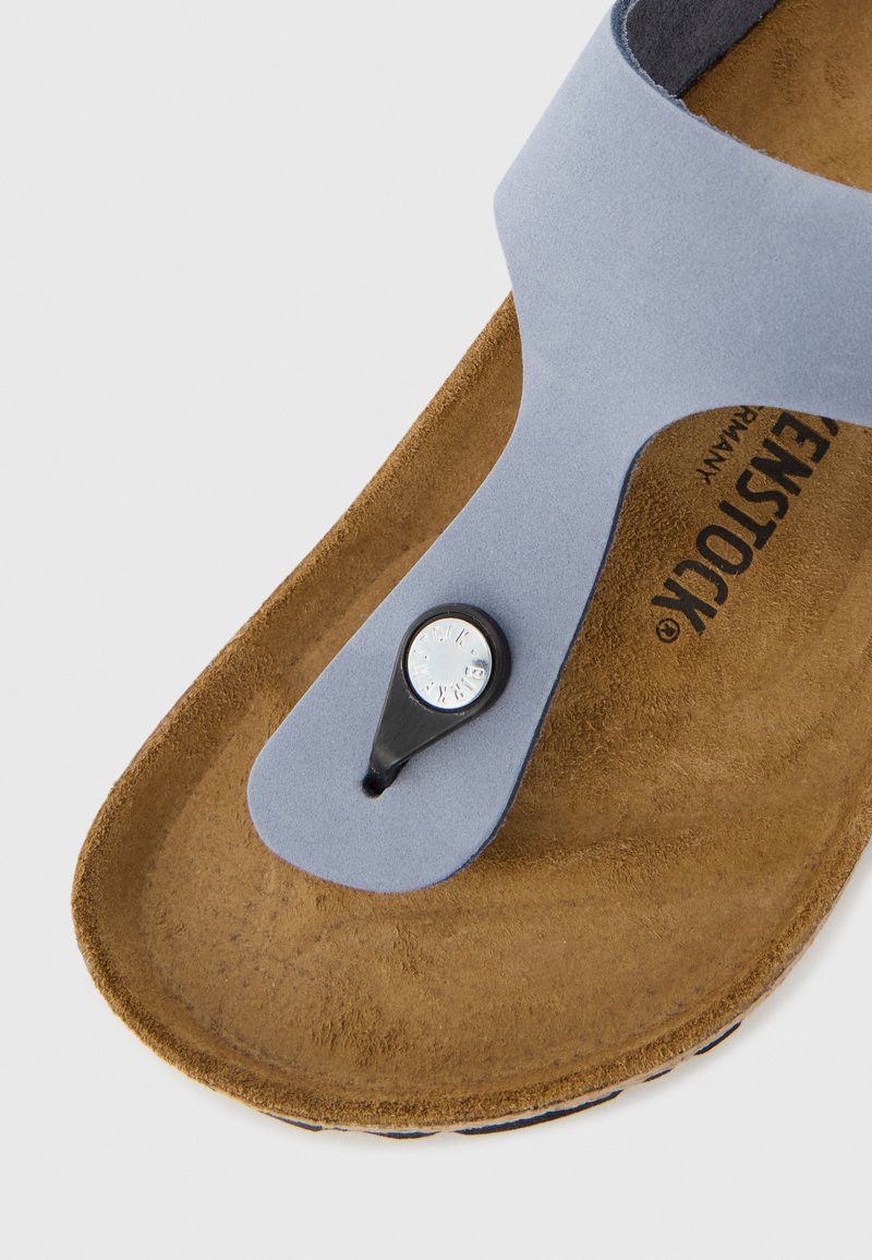 Graue synthetische Sandale mit einem Zehenstegdesign, das mit einem kreisförmigen schwarzen Akzent und einer strukturierten braunen Fußbett ausgestattet ist.