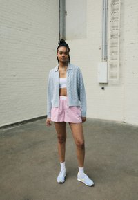 Cardigan de punto trenzado gris claro, top corto blanco, pantalones cortos a rayas rosas y zapatillas azul con acentos amarillos; de pie sobre un suelo de concreto.