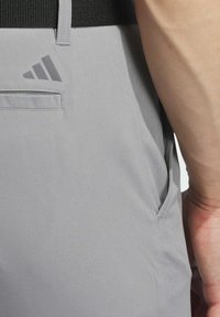 Pantalones cortos deportivos de color gris claro con una tela texturizada, que cuentan con un bolsillo trasero y una cinturilla elástica negra. El logo de Adidas está sutilmente exhibido.