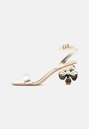 Guldmetallic sandal med åben tå, ankelrem og unik skulptureret hæl formet som en blomst på en lys baggrund.