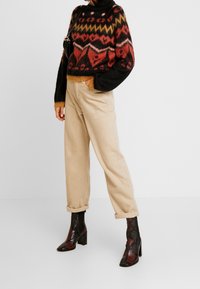 Pull en tricot avec un motif rouge, noir et jaune, assorti à un pantalon beige clair retroussé et des bottines à talons carrés noires et rouges.