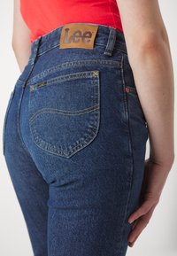 Modré džínsy s vysokým pásom, s koženým štítkom Lee, žltým prešívaním a dvomi zadnými vreckami s krivkovými švami.