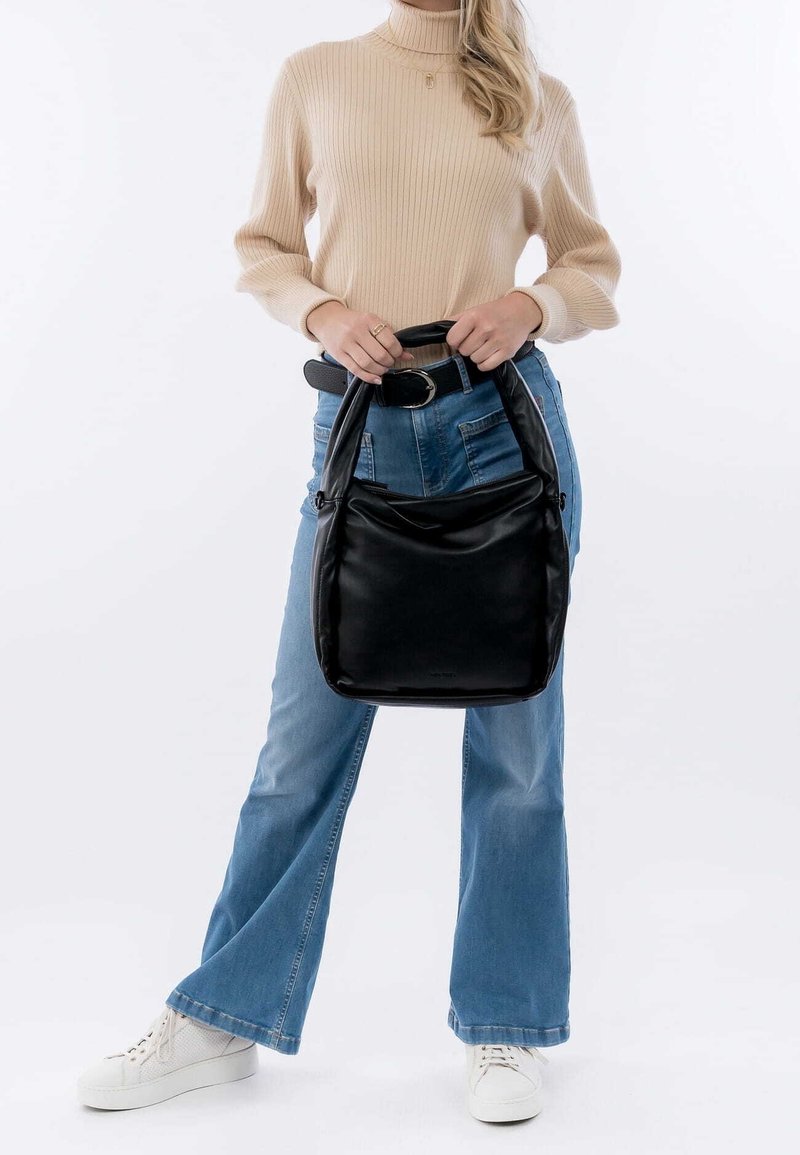 Musta nahkakäekott pehmea, struktureeritud disainiga ja ümarate käepidemetega. Mudel kannab helesiniseid laiad pükse ja valgeid tossusid.