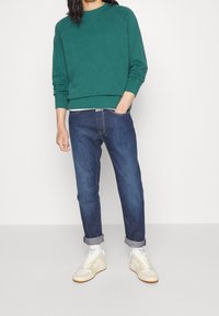 Grön bomullströja med ribbade ärmslut, matchat med mörkblå tapered jeans och vita sneakers med beige detaljer.