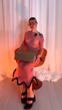 Rochie roz cu dungi și franjuri negri la tiv, purtată cu pantofi platformă negri. Persoana ține o cutie maro marcată cu "MILSTA".