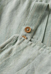 La tela de lino verde claro presenta rayas estrechas y dos botones de madera con un diseño floral. Incluye una tapeta oculta y una cinturilla elástica.