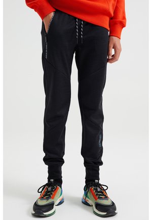 Mustat collegehousut, joissa on vinot saumat, nauhavyötärö ja sivutaskut. Yhdistetty monivärisiin kenkiin, joissa on sekoitus tekstuureja ja kuvioita.