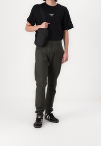 Mężczyzna w czarnej koszulce, oliwkowozielonych spodniach, czarnej torbie na ramię i czarnych trampkach z białymi paskami, stojący na białym tle.