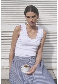Top bianco senza maniche con scollo a volant, abbinato a una gonna a righe. La modella tiene una piccola tazza di caffè su un piatto, con uno sfondo testurizzato.