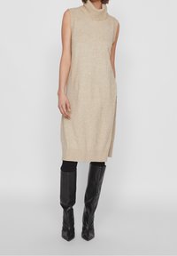 Stickad ärmlös klänning i beige med en hög ribbad krage, med rak passform och gottspringor på sidorna, tillsammans med svarta knähöga stövlar.