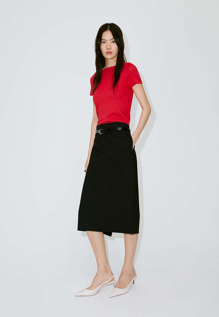 Femme debout portant un haut rouge à manches courtes, une jupe midi noire avec ceinture et des escarpins blancs à bout pointu avec bride arrière sur un fond clair uni.