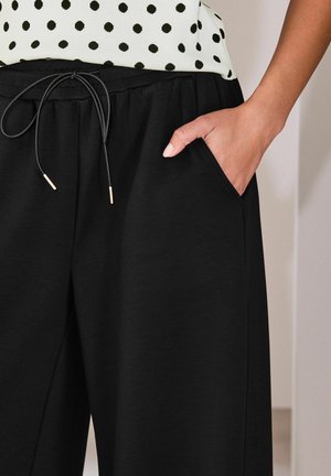 Person trägt schwarze Jogginghosen mit Kordelzug, die Hand in der Tasche, und ein weißes Oberteil mit Punkten, das an der Taille sichtbar ist.