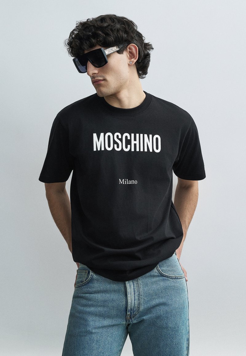 Juoda medvilninė marškinėliai su ryškiu baltu užrašu „MOSCHINO“ ir mažesniu užrašu „Milano“ po juo. Dėvimi su mėlynais džinsais ir dideliais saulės akiniais.