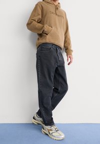 Mies pukeutuneena beigenväriseen Carharttin huppariin, mustiin rentoihin farkkuihin ja beigen ja harmaan sävyisiin lenkkareihin, seisomassa sinisellä matolla valkoista seinää vasten.