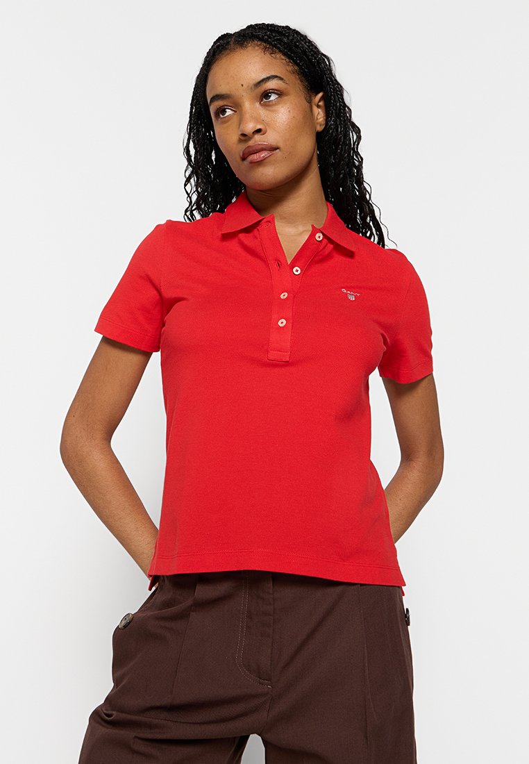 Mujer con cabello rizado vestida con una camiseta polo roja de mangas cortas y pantalones marrones, de pie con las manos en los bolsillos, mirando hacia arriba.