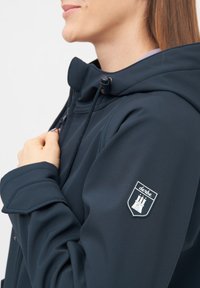 Giacca softshell navy con colletto alto, chiusura con zip e cappuccio regolabile. Presenta un logo ricamato sulla manica destra. Tessuto liscio.