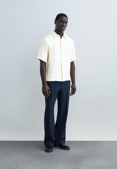 Άνδρας που στέκεται μπροστά σε γκρίζο τοίχο, φοράει κοντομάνικο κρεμ πουκάμισο, σκούρο μπλε παντελόνι και μαύρα παπούτσια, κοιτάζοντας μπροστά με ουδέτερη έκφραση.