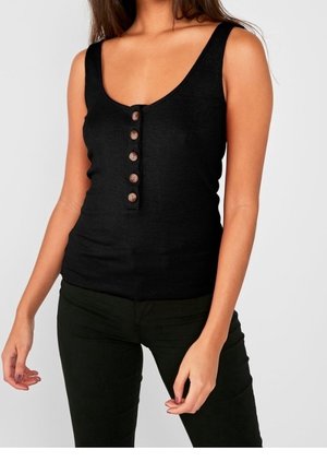 Kvinde iført en sort ærmeløs ribstrikket tanktop med fem brune knapper ned foran, kombineret med sorte bukser.
