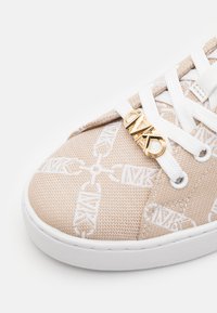 Beige kengät, jotka on valmistettu teksturoidusta kankaasta, valkoisella kuvioidulla designilla ja valkoisilla nauhoilla. Kultaiset yksityiskohdat korostavat nauhoitusaluetta.