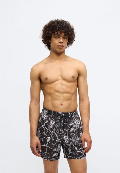 Mladý muž s kučeravými vlasmi, oblečený v čiernych plavkách s bielym kvetinovým vzorom, stojí na pozadí s jednofarebným svetlým pozadím.