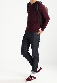 Osoba ubrana w bordowy sweter, ciemne podwinięte dżinsy, białe skarpetki, czerwono-białe trampki i nosząca brązowy plecak na jednym ramieniu.