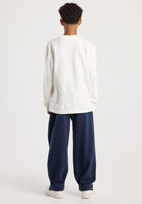 Lapsi, jolla on kiharat hiukset, pukeutunut valkoiseen pitkähihaiseen paitaan, löysiin tummansinisiin housuihin ja valkoisiin tennareihin, seisoo selkä kameraan valkoista taustaa vasten.