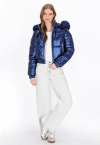 Námořnická modrá lesklá péřová bunda s crop designem, kapucí s kožešinovým lemováním a zipovými kapsami, doplněná o světlé džíny a stříbrné tenisky.