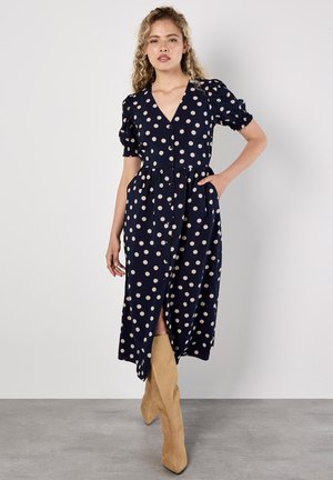 Marineblaues Polka-Dot-Kleid mit kurzen Puffärmeln, V-Ausschnitt und durchgehender Knopfleiste. Merkmale sind eine geraffte Taille und seitliche Taschen sowie ein Schintz bis zum Oberschenkel.